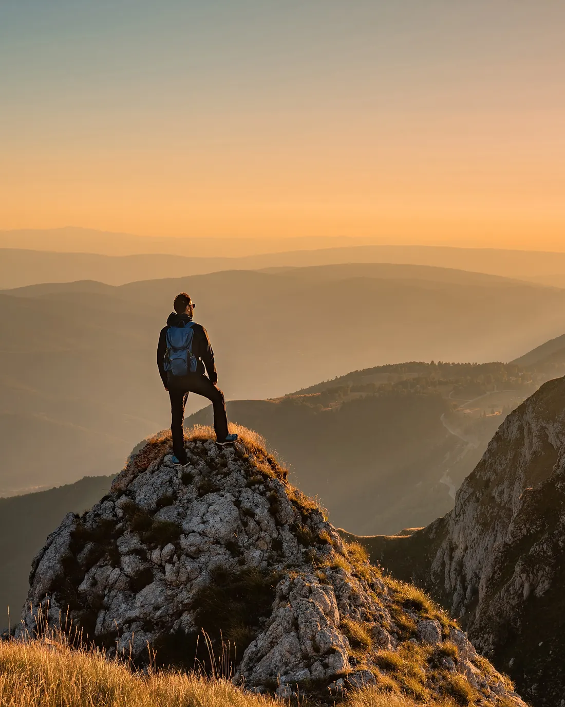 A hiker standing on a mountain summit at sunset, looking out over a wide valley